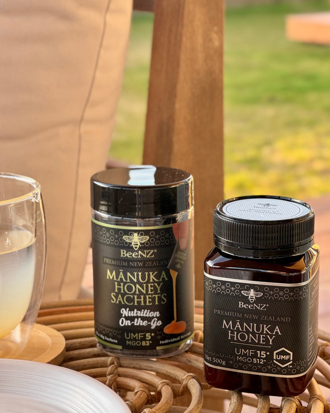 Two jars of BeeNZ Manuka Honey on a table with a blurred outdoor background