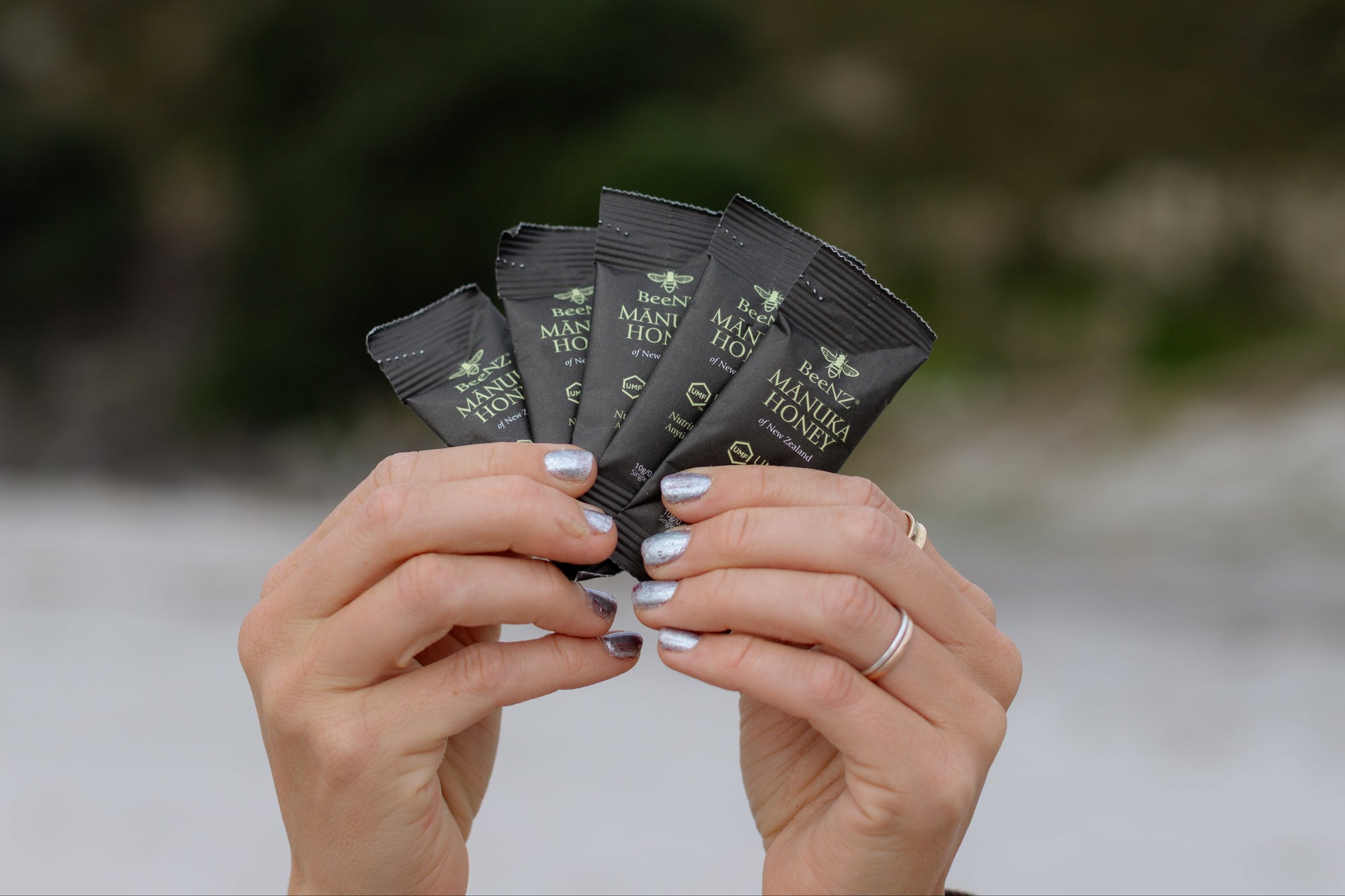 A person's hands holding a fan of four black manuka honey sachets against a blurred outdoor background of a grassy hill and water.