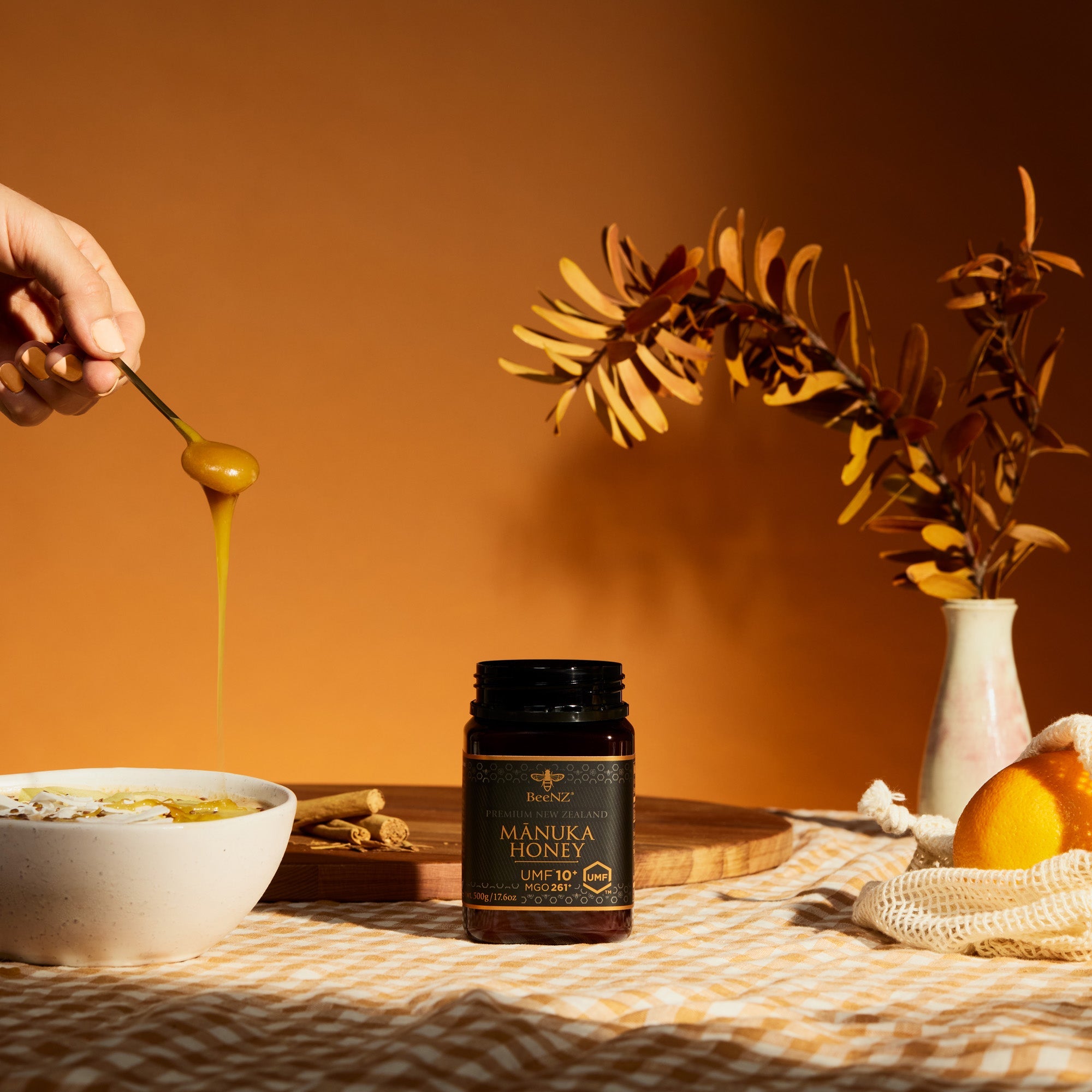 UMF10+ MGO 261 BeeNZ Manuka honey being spooned into a bowl with a jar of Manuka Honey on a warm-toned tablecloth. 