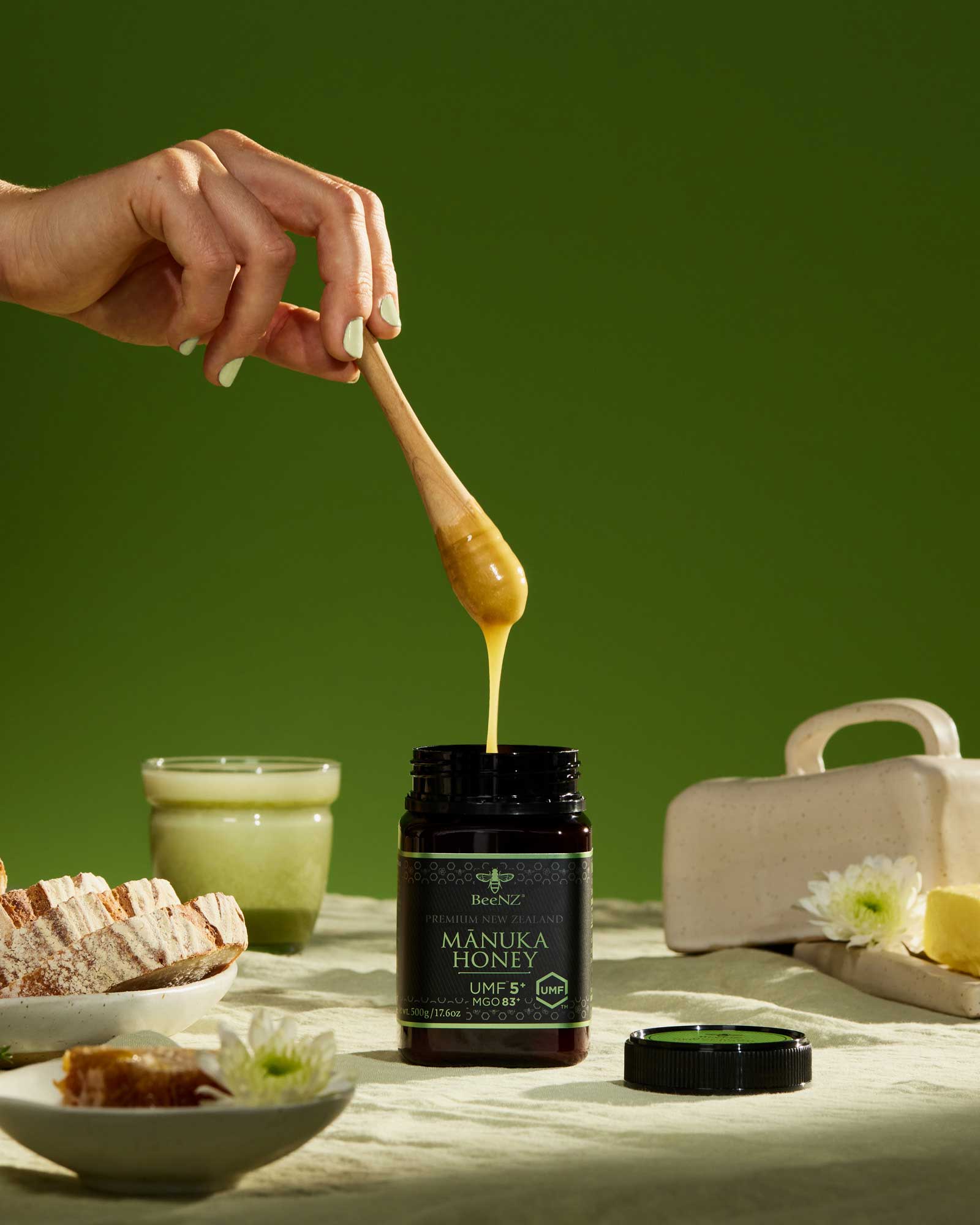 A woman's hand holding a wooden honey stirrer with premium New Zealand BeeNZ Manuka Honey dripping into the honey jar. Honey jar is on a table with a linen tablecloth and sourdough and butter on plates.